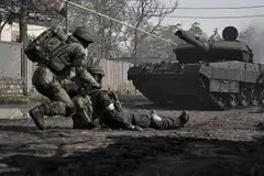 A soldier in Wardogs dragging a fallen ally across the road as a tank advances toward them.
