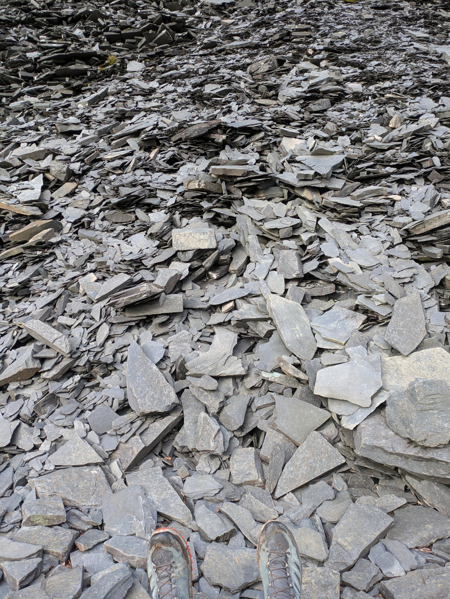 A huge pile of broken slate, filling the whole picture, with a pair of booted feet at the bottom.
