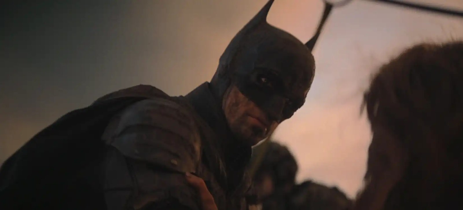 A very grubby-looking Batman (Robert Pattinson) looks down at a prone woman he’s rescuing in The Batman